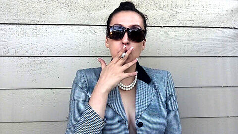 Affascinante donna d'affari con la collana di perle e la manicure alla francese che fuma una sigaretta bianca a filtro 100 in pubblico, mostrando il suo decoltè e le sue tette sode attraverso gli occhiali da sole.